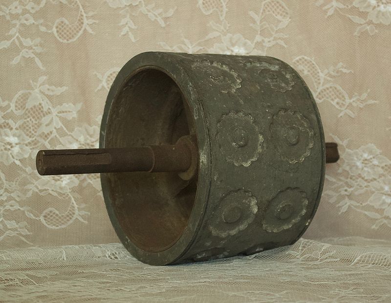 Zijaanzicht van een antieke metalen deegroller met verhoogde rozetpatronen en een lange metalen as