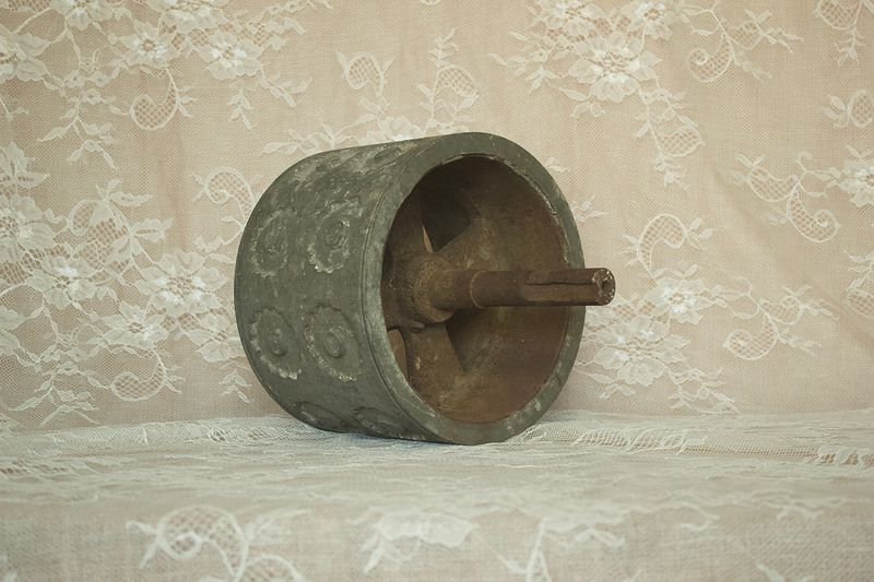 Zijaanzicht van een antieke metalen deegroller met verhoogde rozetpatronen en een lange metalen as