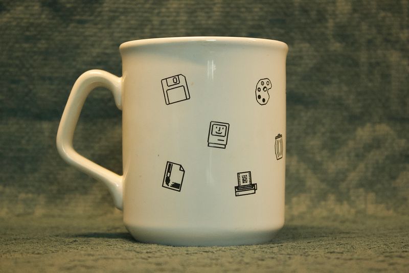 Achteraanzicht van de vintage Apple-mok, waarop iconen te zien zijn van een floppy disk, een verfpalet, een prullenbak 